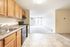 Bright open-plan living space with wooden cabinetry, modern appliances in the kitchen, and a spacious carpeted living area leading to a sunlit balcony.