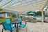 Princeton Westford Apartments | Outdoor patio area at Princeton Westford Apartments features a round black table and four bright blue chairs under a pergola, with a community grilling area and fenced swimming pool with blue umbrellas and lounge chairs nearby. | Wheelchair Accessible Apartments in Westford