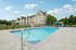 Princeton Westford Apartments | Outdoor salt water pool with metal handrail, surrounded by lounge chairs and umbrellas. Behind the pool, Princeton Westford Apartments rise above a grassy lawn and trees under a blue sky with scattered clouds. | Apartments with Balcony in Westford