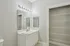 Grandview Apartments | A modern bathroom at Grandview Apartments in Lowell, MA features a white vanity, sink, and mirror under bright lights next to an open closet with white wire shelves, set against light gray walls and a beige tiled floor. | Wheelchair Accessible Apartments in Lowell