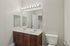 Grandview Apartments | A modern bathroom at Grandview Apartments in Lowell, MA features a double-sink vanity, large mirror with lights above, wooden cabinets, and a white toilet. Light-colored walls and a slightly open door complete the space. | Pet Friendly Apartments in Lowell