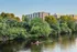 A person rows a red single scull boat on a calm river, surrounded by green trees with The Charley Apartments in Allston MA visible in the background under a clear blue sky. | The Charley | Apartment in Allston, MA