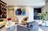 A modern living room at The Charley Apartments in Allston MA features patterned and yellow armchairs, a white oval coffee table with flowers and books, built-in lit wall shelves, abstract art, a kitchenette, and a green indoor plant | The Charley | Apartment in Allston, MA