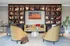 A modern living room at The Charley Apartments in Allston MA features four cushioned chairs around a white coffee table, bookshelves along the back wall, a TV showing a tennis match, a brown handbag on the floor, and a potted plant to the right. | The Charley | Apartment in Allston, MA