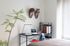 A minimalist home office at The Charley Apartments in Allston MA features a small desk, laptop, books, and lamp beside a shelf of files. Two wooden masks hang above, while a potted plant and sunlight add natural elements to the cozy room. | The Charley | Apartment in Allston, MA