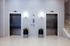 Two stainless steel elevator doors are set in a light-colored wall at Charley Apartments in Allston MA. Each elevator has a button panel and a potted plant in front, with a staircase to the right. The floor is tiled. | The Charley | Apartment in Allston, MA
