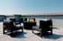 A modern rooftop patio at Charley Apartments in Allston MA features four black cushioned chairs around a round table, with potted plants and blue sky in the background. | The Charley | Apartment in Allston, MA