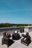 A rooftop patio at Charley Apartments in Allston MA features five black cushioned chairs around a round fire pit table, decorative pillows, potted plants, and a scenic view of trees, the river, and distant buildings. | The Charley | Apartment in Allston, MA