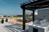 A modern rooftop patio at Charley Apartments in Allston MA features a pergola, outdoor kitchen, grill, yellow chairs and tables, potted plants, and a large TV screen showing a soccer game against the city skyline backdrop. | The Charley | Apartment in Allston, MA