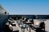 Modern rooftop patio at Charley Apartments in Allston MA with black lounge chairs, small tables, potted plants, and decorative pillows, set against a clear blue sky and minimalistic railing. Two lounge chairs are in the background. |The Charley | Apartment in Allston, MA