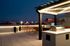 A modern rooftop terrace at Charley Apartments in Allston MA features a lit pergola with a built-in grill, bar-height tables and chairs, city buildings in the background, and potted plants along a sleek railing. | The Charley | Apartment in Allston, MA