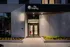 The entrance to The Charley Apartments in Allston MA features a modern design with the sign The Charley above glass doors, the number 46 on the wall, plants along the walkway, and exterior lights illuminating the area at dusk. | The Charley | Apartment in Allston, MA