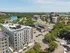 Aerial view of The Charley Apartments in Allston MA, surrounded by trees, with a river on the left, nearby roads, and urban buildings in the background under a clear blue sky. | The Charley | Apartment in Allston, MA