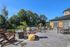 An outdoor patio at The James Apartments in Haverhill, MA features wooden chairs, tables, and a fire pit, all surrounded by greenery and a fence. Yellow patterned ottomans bask in bright sunlight under a clear blue sky. | The James | Apartment in Haverhill, MA