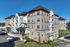 The James Apartments in Haverhill MA is a modern four-story building with gray and tan siding, large windows, and stone accents, surrounded by landscaped areas and parked cars under a clear blue sky. | The James | Apartment in Haverhill, MA