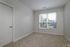 An empty room at The James Apartments in Haverhill, MA features beige carpet, light gray walls, a white door, and a large window with white blinds letting in natural light. Trees and neighboring buildings are visible outside the window.  | The James | Apartment in, Haverhill MA