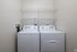 A laundry room at The James Apartments in Haverhill MA features a white washing machine and dryer side by side against a beige wall, below two empty white wire shelves. | The James | Apartment in, Haverhill MA