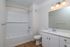 Modern bathroom at The James Apartments in Haverhill MA featuring white walls, a bathtub with white subway tiles, a toilet, sleek vanity with sink, large mirror, and three-light fixture above. Wood-like flooring completes the contemporary design.  | The James | Apartment in, Haverhill MA