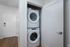 A stacked white washer and dryer set is installed in a small laundry closet with white walls and a partially open door, located in the hallway of The James Apartments in Haverhill MA, featuring modern wood flooring.  | The James | Apartment in, Haverhill MA