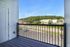 A view from a balcony with black railings at The James Apartments in Haverhill, MA, overlooking a parking lot, road, and green buildings set against a tree-covered hill under a clear blue sky.  | The James | Apartment in, Haverhill MA
