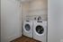 A laundry closet at The James Apartments in Haverhill, MA features a front-loading washer and dryer side by side beneath two white wire shelves, set against a neutral wall and wood flooring. The closet door is partially open. | The James | Apartment in, Haverhill MA
