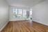 A bright, empty room at The James Apartments in Haverhill MA features light-colored walls, large windows with blinds, a glass door to the patio, and wood flooring. Natural light fills the space with views of a grassy outdoor area.  | The James | Apartment in, Haverhill MA