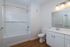 A modern bathroom at The James Apartments in Haverhill, MA, features a white bathtub and shower, a toilet, a sleek vanity with sink and mirror, three lights above the mirror, wood-style flooring, and light beige walls.  | The James | Apartment in, Haverhill MA