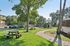 Pheasant Run Apartments | A landscaped building entrance at Pheasant Run Apartments in Nashua NH features colorful flower beds, neatly trimmed shrubs, a large tree, and a blank blue sign on a lawn along a curved sidewalk. | Nashua Apartments with Virtual Tours