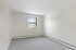 Princeton Properties | A small, empty room at Brookstone Manor Apartments in Milford, New Hampshire features light gray walls, a single window letting in daylight, gray carpeted floor, and white baseboard heating along the walls. | Apartments with Air Conditioning in Milford