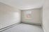 Princeton Properties | Empty room at Brookstone Manor Apartments in Milford, New Hampshire, featuring light gray walls, a white ceiling, gray carpet, baseboard heaters, and a single window letting in natural light. | Apartments with Air Conditioning in Milford