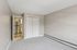 Princeton Properties | Empty room at Brookstone Manor Apartments in Milford, New Hampshire, featuring beige walls, white trim, gray carpet, a closed white closet, and an open door to a kitchen with wood flooring and a stainless steel refrigerator. | Milford Apartments with Virtual Tours