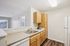 Bright and modern kitchen with wooden cabinets, sleek countertops, and a built-in dishwasher, offering a glimpse into a sunlit living area with large windows.| Village Green | Apartment in Tewksbury, MA