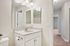 Modern bathroom with sleek white vanity and large mirror, bright lighting, and neutral walls leading to the hallway.| Village Green | Apartment in Tewksbury, MA