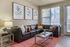 Modern living room with a sleek black leather sectional, vibrant orange rug, and stylish botanical artwork. Natural light streams through large windows, creating a warm and inviting atmosphere.