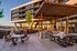 Modern apartment building with a vibrant outdoor social area, featuring sleek patio furniture, a BBQ station, and relaxed gatherings under a wooden pergola during sunset.