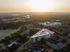 Aerial view of a modern triangular apartment building bathed in warm sunrise light, surrounded by lush greenery and quiet streets, offering a serene urban living experience.