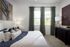 Elegant bedroom with stylish decor, plush pillows on a cozy bed, large windows with green curtains, and modern art accents.