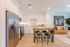 Modern open kitchen and living area featuring elegant stainless steel appliances, a chic wooden dining table with stylish bar stools, and a cozy leather couch accented by vibrant artwork and greenery.