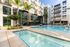 Luxurious apartment complex featuring a sunlit courtyard with a sparkling swimming pool and modern lounge chairs, framed by tall palm trees and elegant architecture.