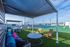 Modern rooftop terrace with shaded seating, vibrant cushions, and tennis court overlooking cityscape under a clear blue sky.