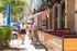 Charming urban scene with people walking on a vibrant street lined with modern buildings, lush greenery, and cozy outdoor seating under warm string lights.