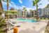 Luxurious apartment complex featuring a pristine pool surrounded by stylish lounge chairs and vibrant green umbrellas, set amidst modern architecture and lush palm trees, under a clear blue sky.