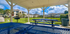 Serene apartment view from a covered patio with blue picnic tables, a barbecue grill, and lush greenery around a tranquil pond.