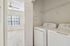 Bright laundry room with modern washer and dryer, adjacent to a sunlit room with carpet flooring and large window.