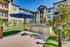Charming apartment complex with modern outdoor grilling area, featuring stainless steel BBQ grills and a spacious patio under a blue umbrella, surrounded by lush greenery.