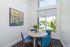 Bright and modern dining nook featuring a round table with vibrant blue chairs, large windows with lush green views, and an artful beach painting adding a touch of serenity.