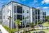 Modern three-story apartment building with sleek balconies and lush landscaping under a bright blue sky.