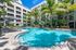 Luxurious apartment complex featuring a stunning palm-fringed swimming pool, surrounded by lush greenery and stylish loungers, under a clear blue sky.