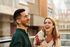 Two people laughing and enjoying a sunny day on a balcony, with urban buildings in the background.