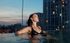 Elegant woman relaxing in a rooftop infinity pool with a cityscape backdrop at sunset.